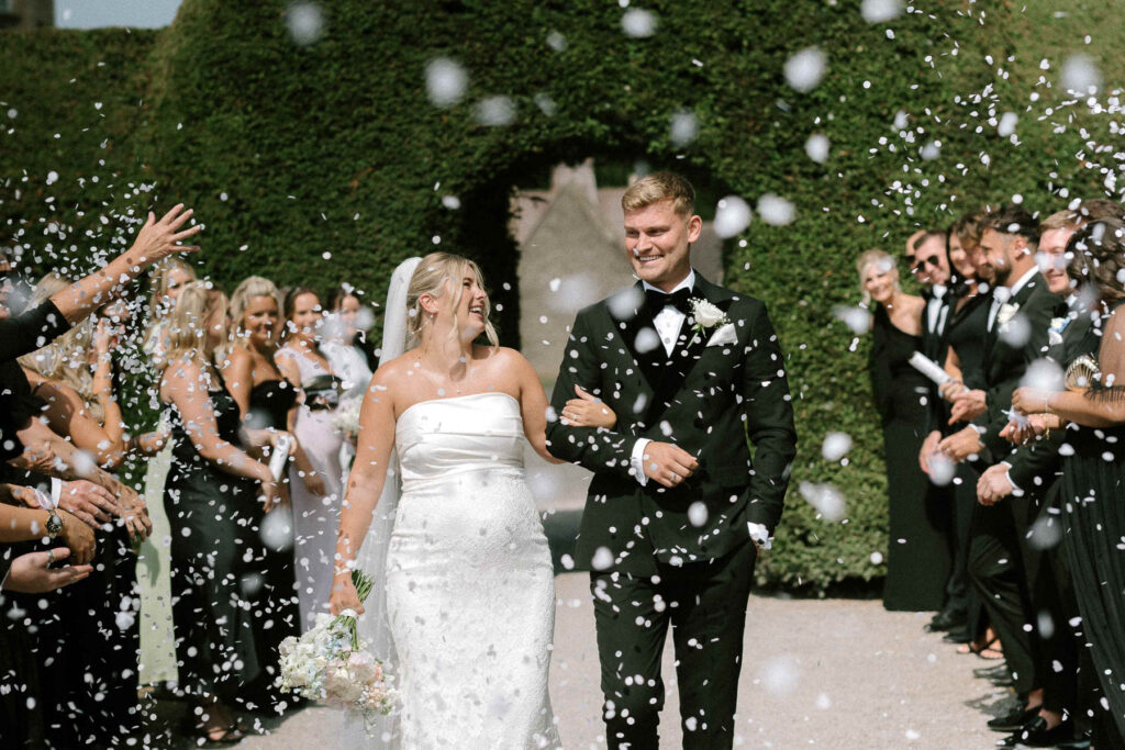 bride and groom walking through confetti tunnel with guests throwing white paper confetti at the couple