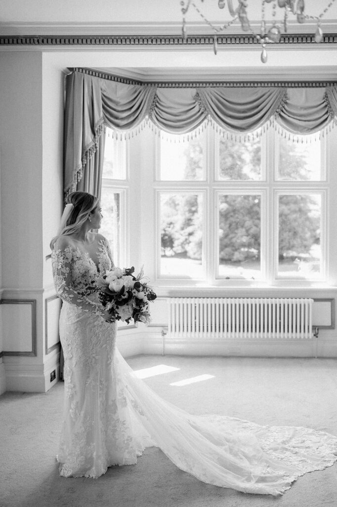 bride wearing lace dress and veil holding bouquet looking out of window on wedding day
