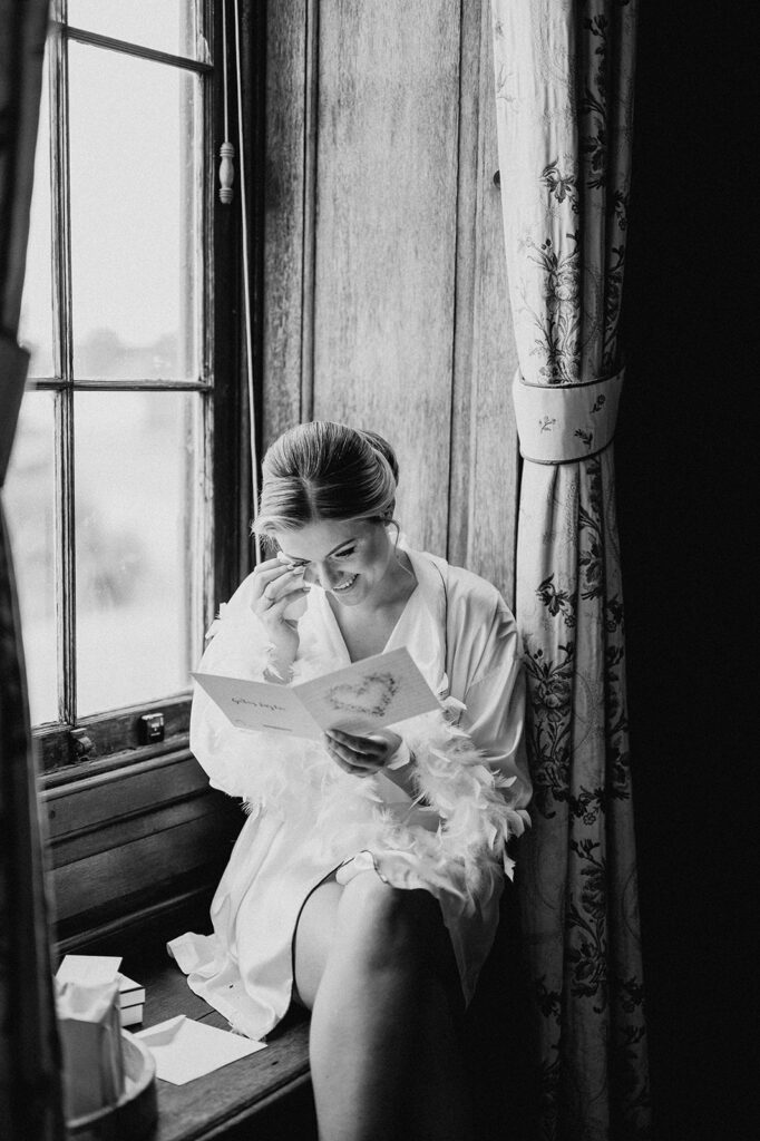 bride opening letter from her groom on her wedding morning sitting on windowsill at glemham hall