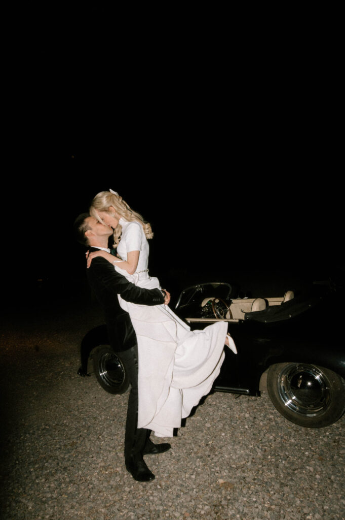 groom lifting bride in front of vintage wedding car