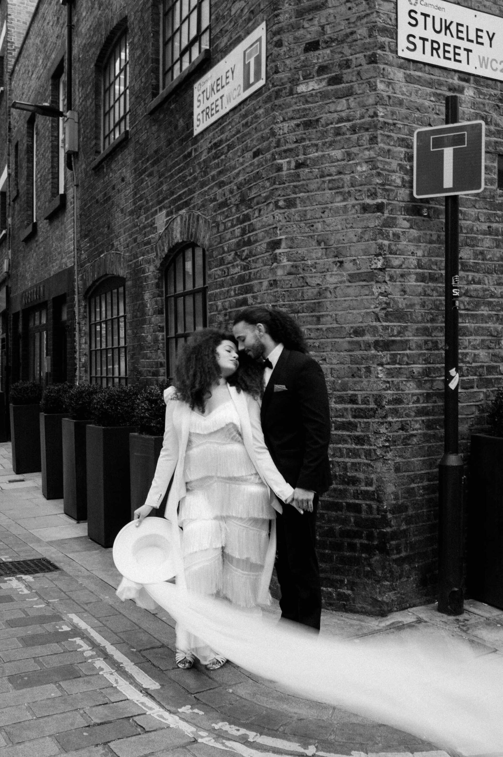 bride wearing white jumpsuit and holding white hat holding hands with groom in streets of London