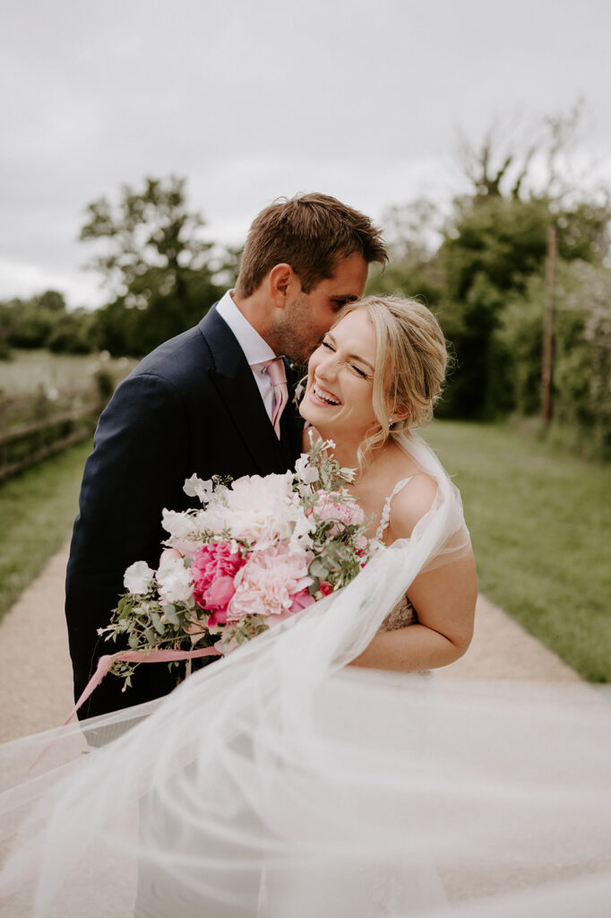 groom kissing bride wearing Martina liana wedding dress at retreat east holding pink peony wedding bouquet