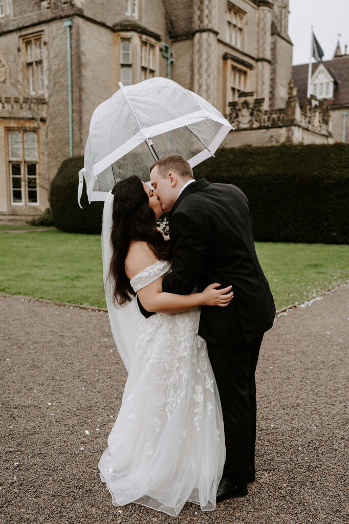 bride and groom sharing a kiss with clear umbrella walking in the rain at tortworth court