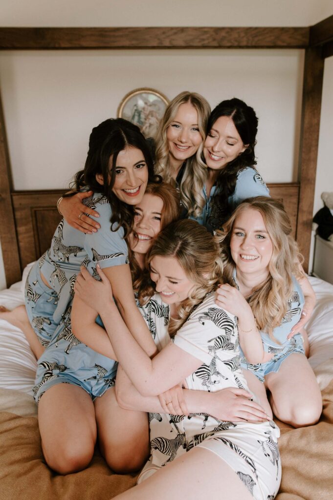 Bridesmaids sitting together hugging on the bed in their robes during bridal prep at Bruisyard Hall