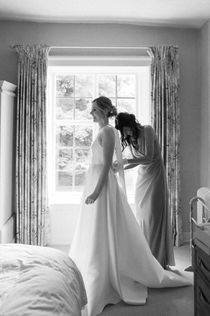 Black and white photo of bride Heidi having her dress done up by the window at Bruisyard Hall