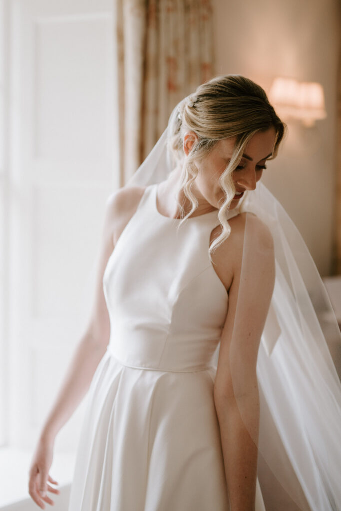 Bride adjusting her wedding dress in soft window light at Bruisyard Hall