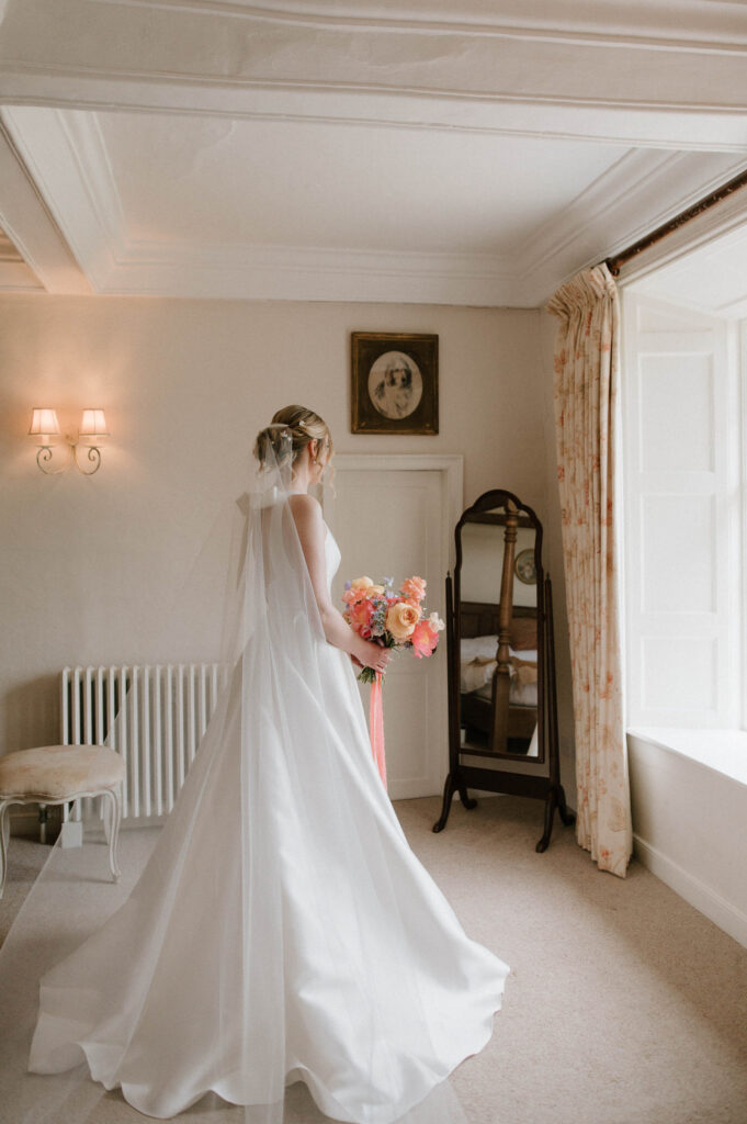 Bride Heidi standing by the window holding her bouquet during bridal prep at Bruisyard Hall