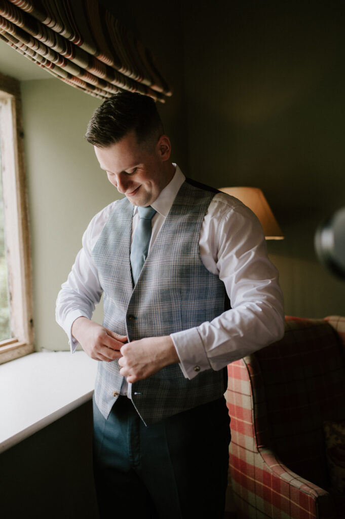 Groom Charley adjusting his waist coat  in the green room during prep at Bruisyard Hall