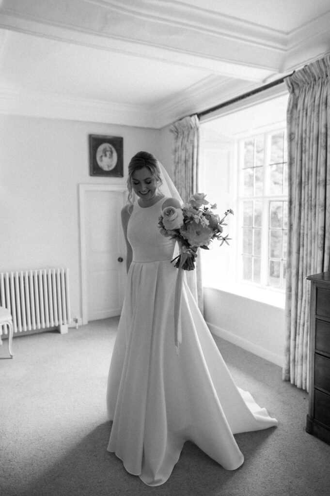 Black and white photo of bride Heidi standing by the window holding her dress during prep at Bruisyard Hall