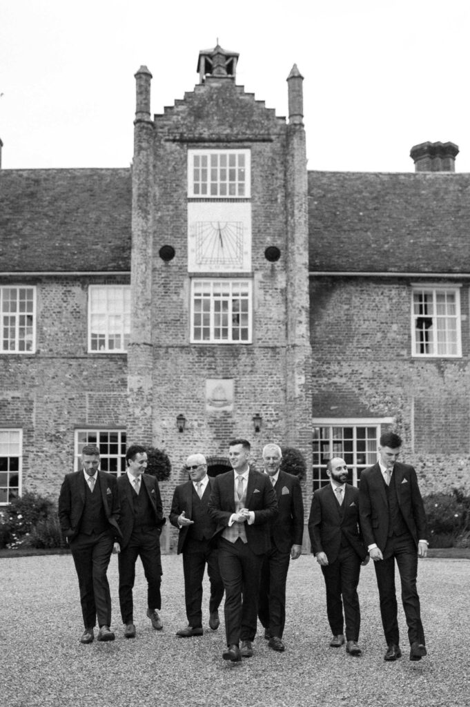 Black and white photo of groomsmen standing and  joking together outside Bruisyard Hall