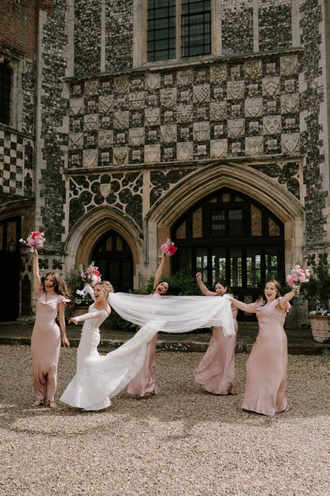 Bridesmaids lifting the bride’s veil outside Butley Priory during bridal party photos.