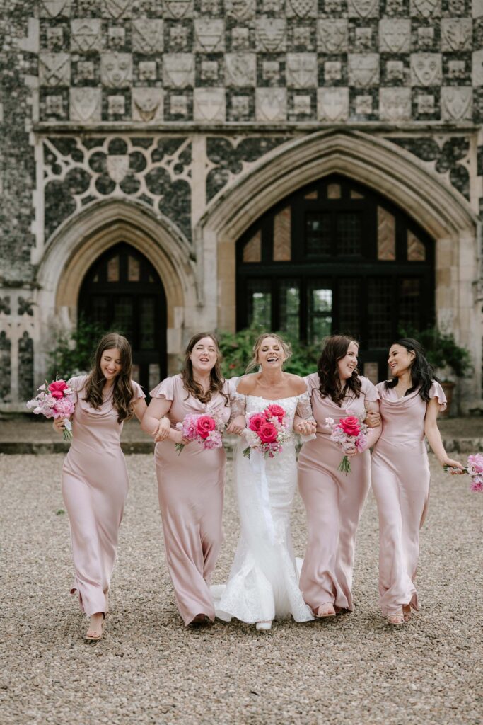 Bride walking with her bridesmaids outside Butley Priory holding pink bouquets.