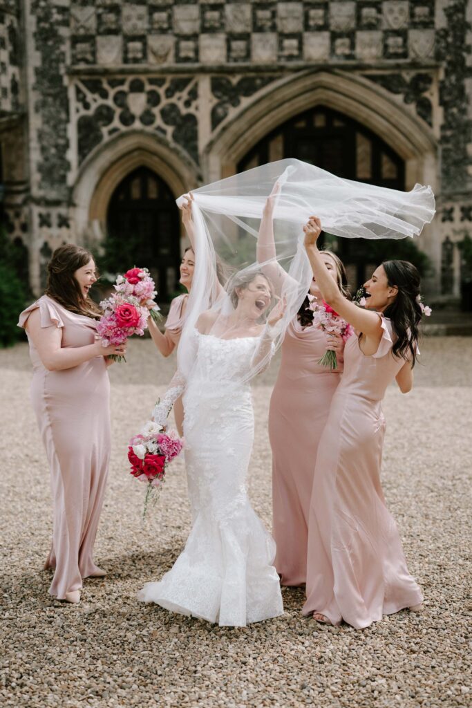 Bridesmaids lifting the bride’s veil during fun bridal party portraits at Butley Priory.

