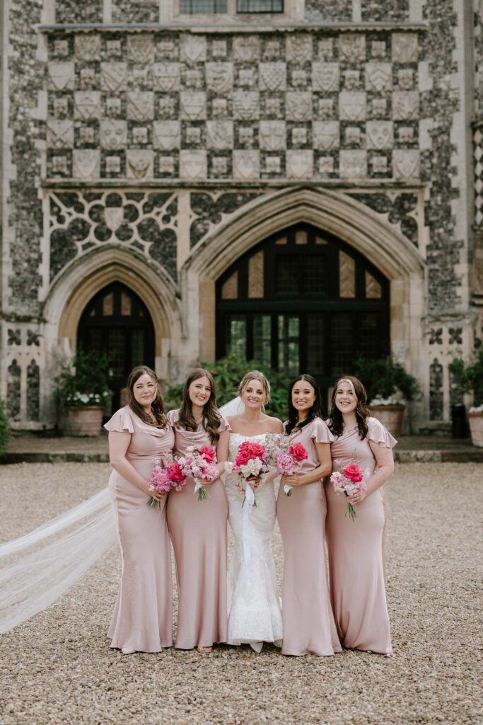 Bridesmaids standing together outside Butley Priory holding pink wedding bouquets.