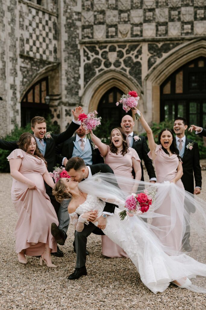 Wedding party cheering as the groom dips the bride playfully outside Butley Priory.