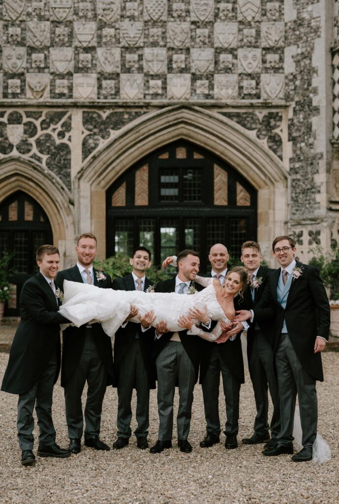 Groomsmen lifting the bride during a fun group portrait outside Butley Priory.
