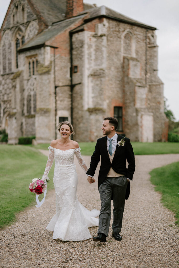 Bride and groom holding hands and laughing together outside Butley Priory.