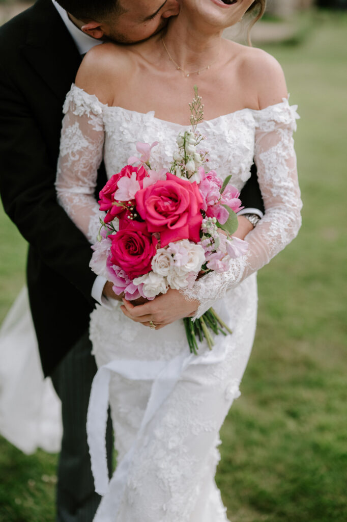 groom holding bride from behind whilst she holds her bright pink and white rose bouquet