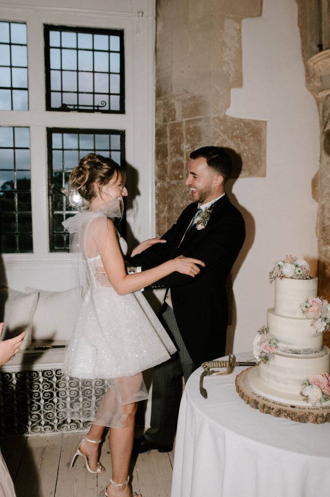 Bride and groom holding hands and smiling during cake cutting.
