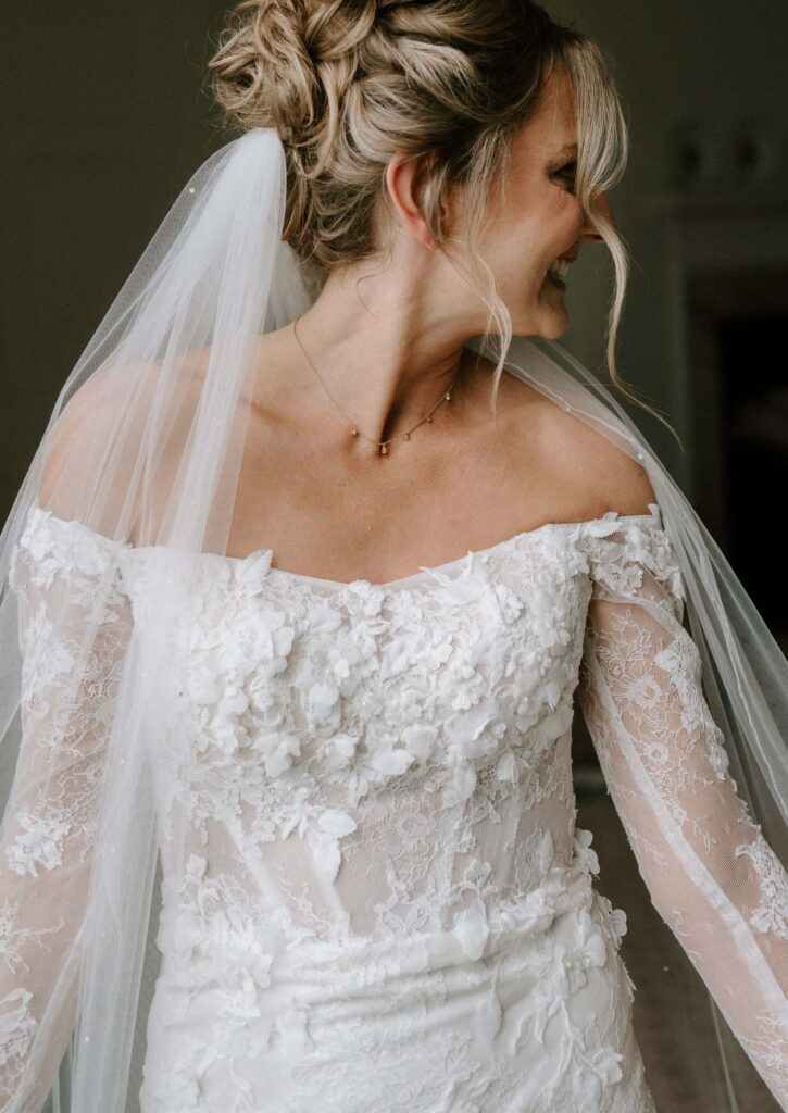 Bride smiling during bridal prep at Butley Priory, wearing an off-the-shoulder lace wedding dress and veil.