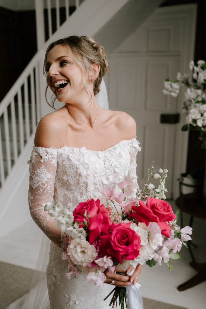 bride wearing off the shoulder white lace wedding dress smiling holding pink and white bouquet at butley priory