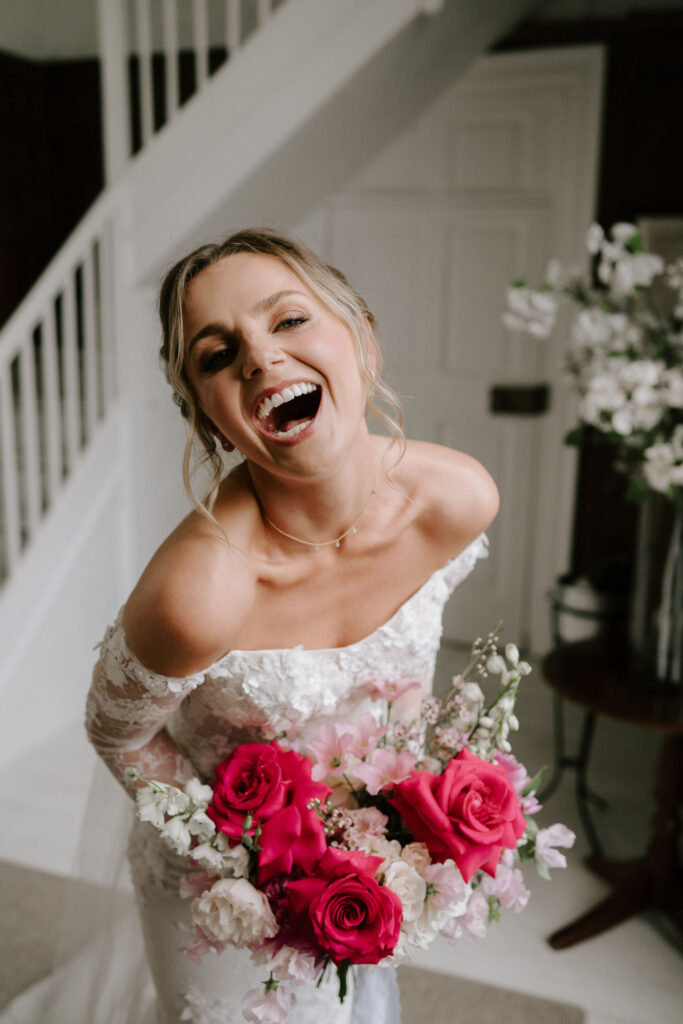 bride wearing off the shoulder white lace wedding dress smiling holding pink and white bouquet at butley priory