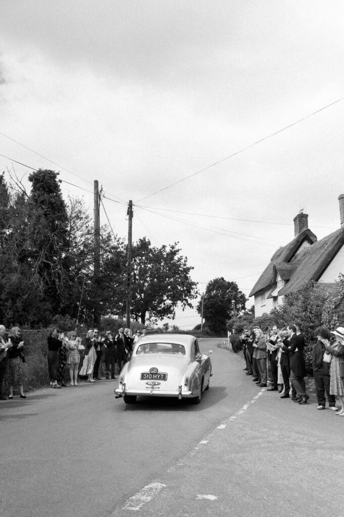 Vintage wedding car driving through a village with guests watching after the ceremony.