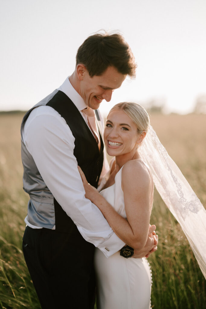 Bride smiling at camera while being held by groom at sunset