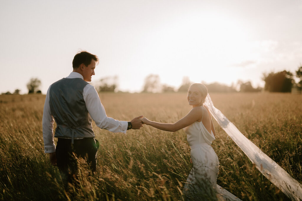 Bride and groom holding hands while walking through long grass at sunset