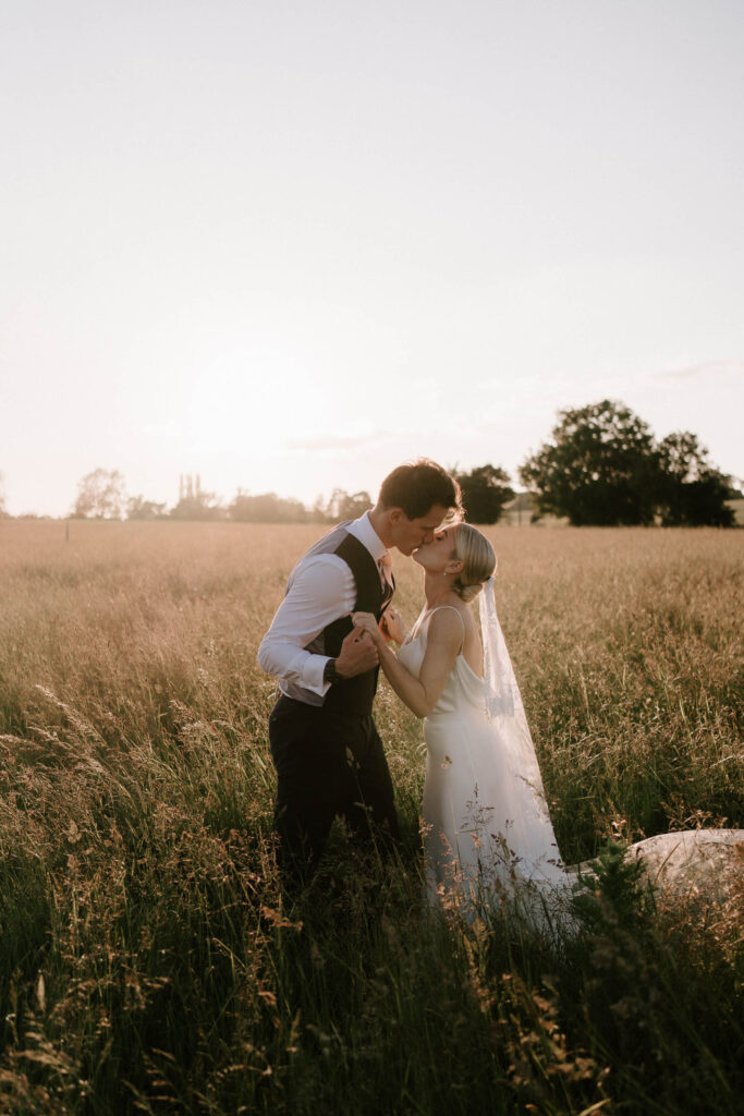 Bride and groom kissing in long grass during golden hour