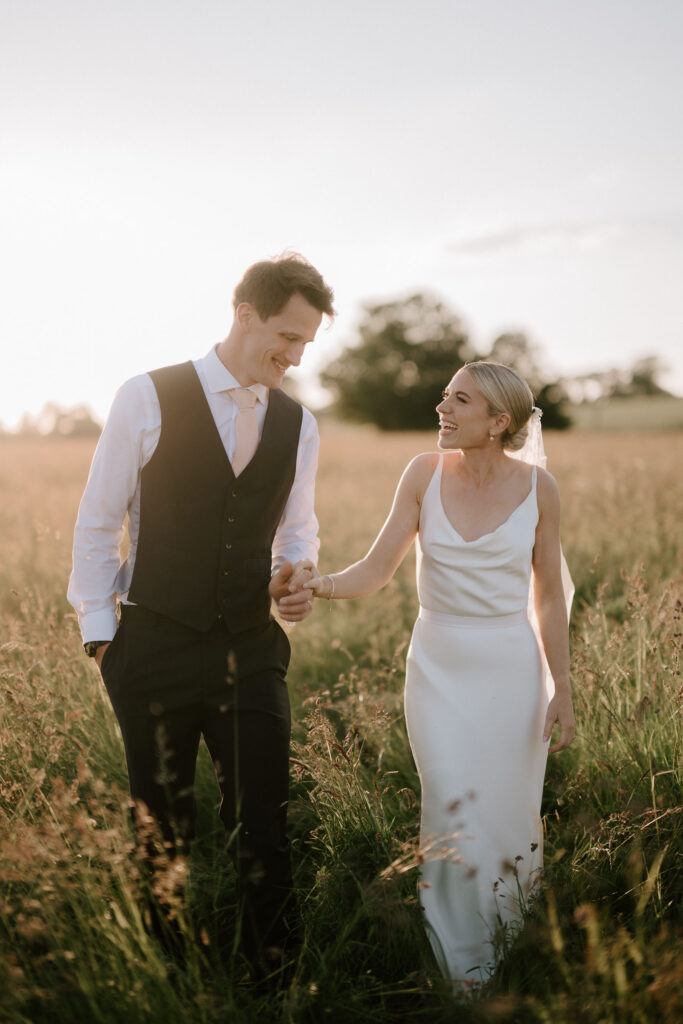 Bride and groom walking through long grass during golden hour