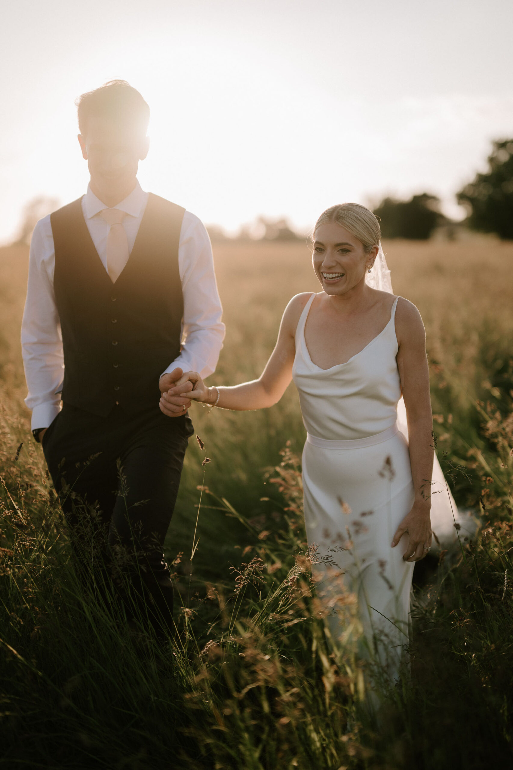 bride and groom walking through field at golden hour in suffolk