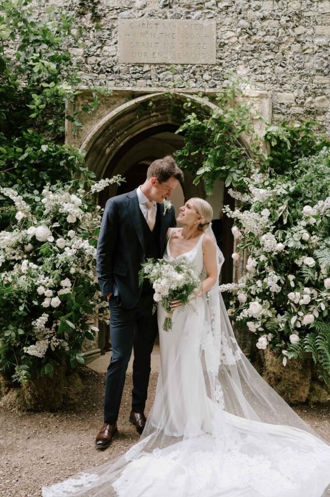 Bride and groom standing together beneath floral arch outside the church in Earl Soham, Suffolk