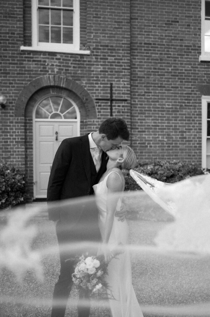 Bride and groom standing close together and sharing a kiss with the veil blowing in the breeze