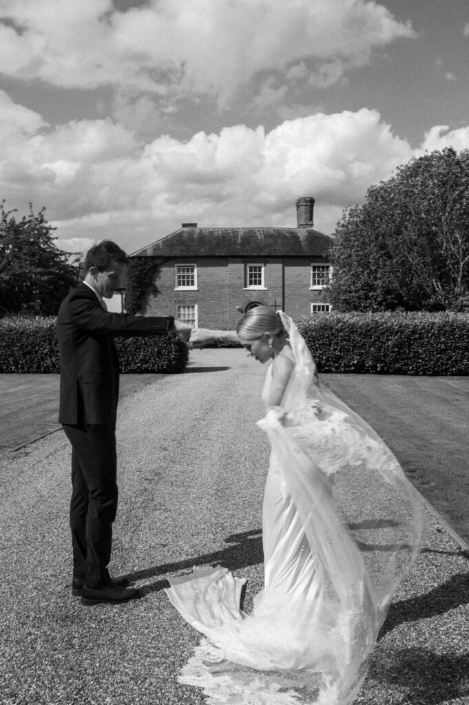 Bride playfully lifting her dress while the groom waits on the gravel driveway